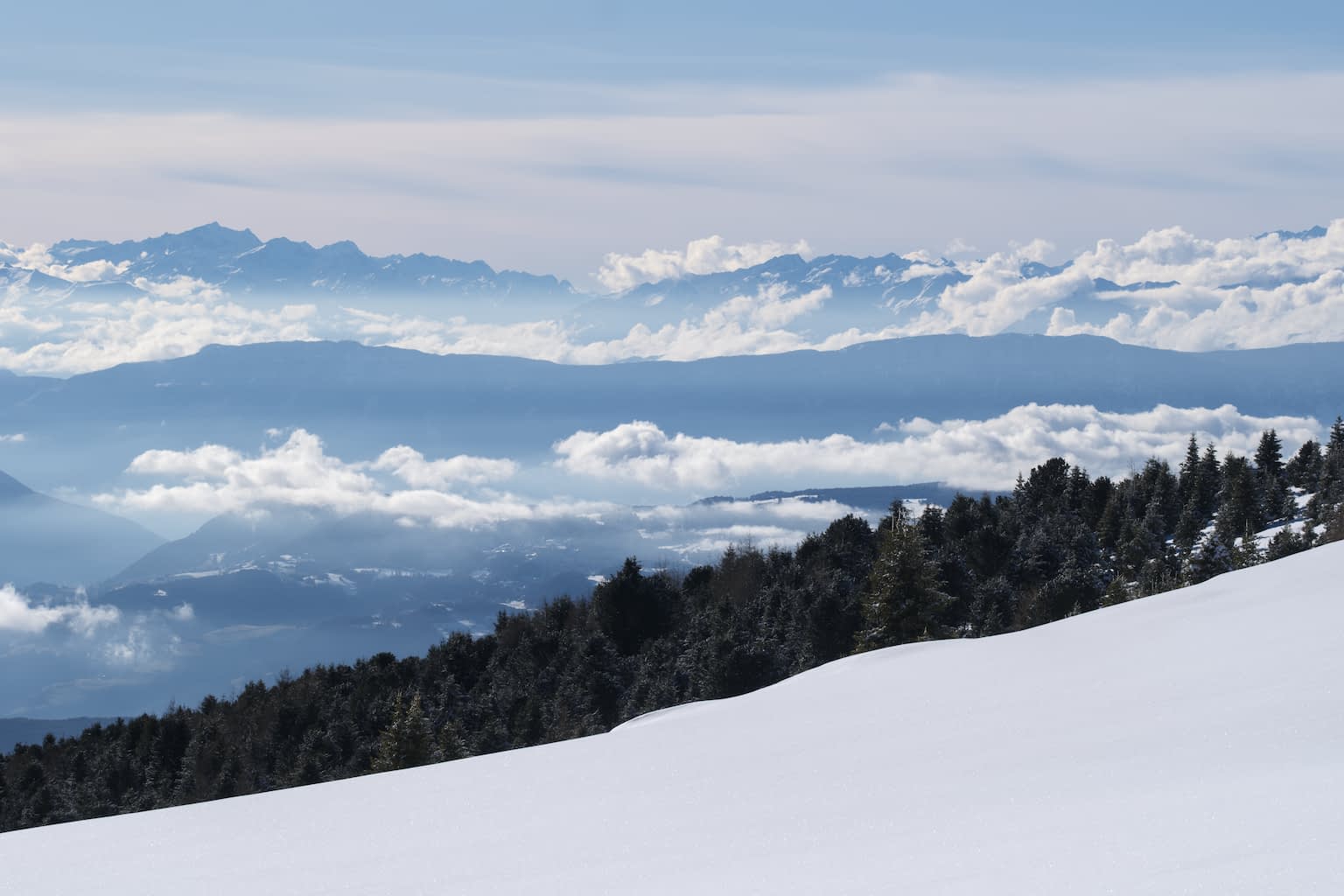 Winder day in the mountains with partially cloudy sky at the top and untouched snow cover at the bottom delineated by a dark stripe of trees and blue valleys filled by fog clouds.