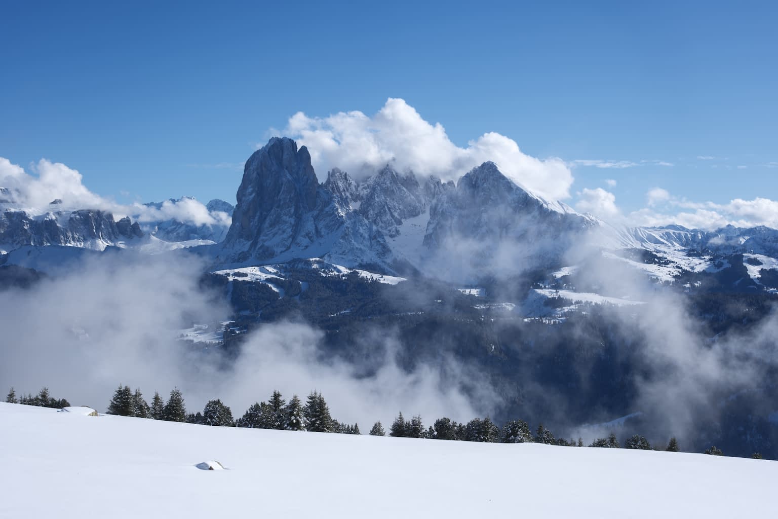 The Langkofel Group in the winter embraced by fog clouds with a clear blue sky above and a snowy hill below