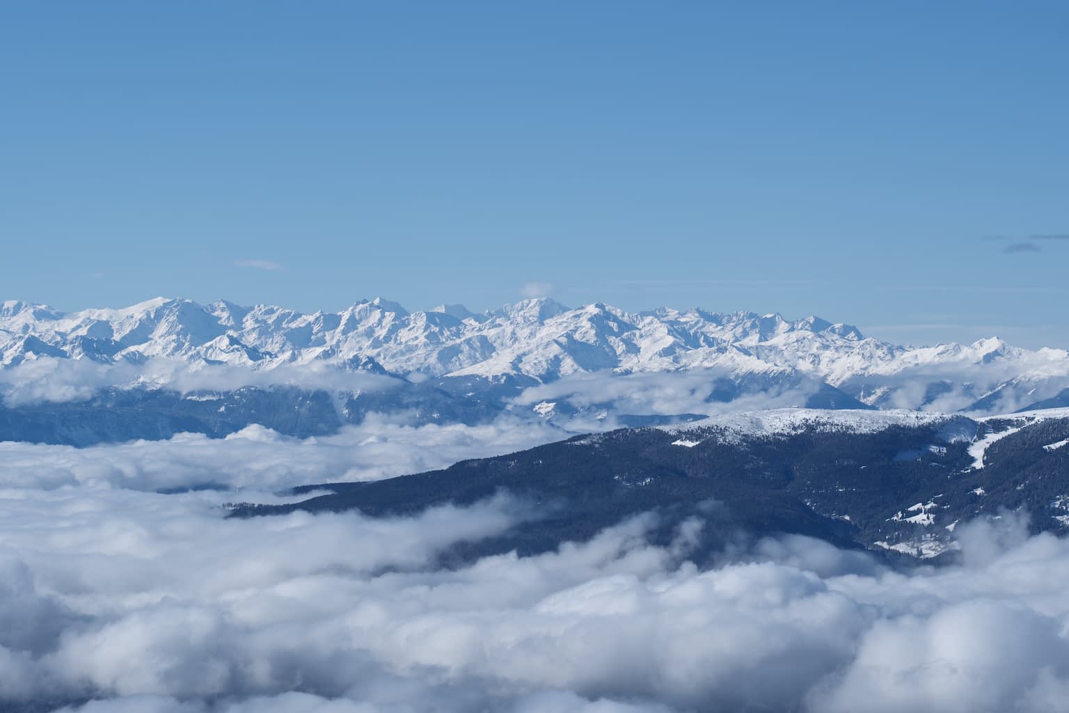 View of a mountain range with the Ortler in the center and the Ritten in the foreground, fog covering the lower fifth and blue sky the upper third.