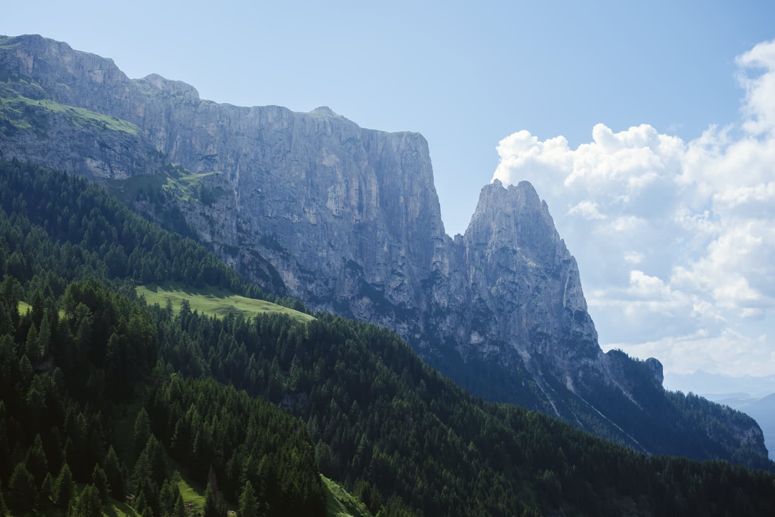 The mountain Schlern as seen from the north colored in a cold blue with fluffy clouds to the right and a clear blue summer sky to the left and green trees filling the bottom left corner.