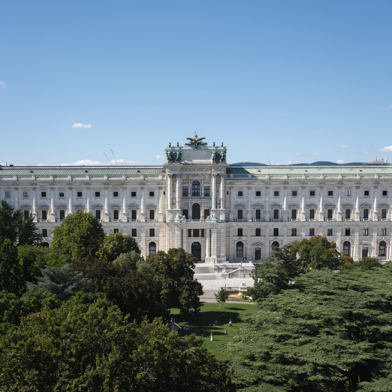 The Hofburg in Vienna as viewed from the Burggarten with a clear blue sky above and the lush green garden in front.