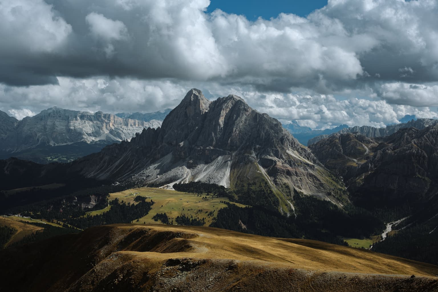 The Peitlerkofel peek in the center in early autumn with an orange-brown foreground and a cyan-blue sky mostly covered by clouds.