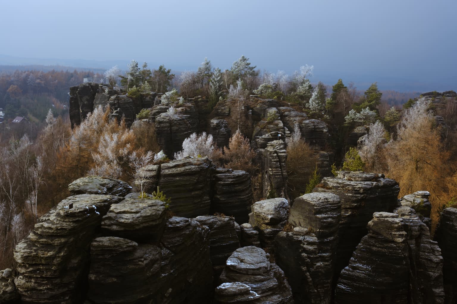 The Tiské stěny rock formation in late autumn with vertical black rock pillars forming groups and trees filling the gaps in between and sitting atop some of the peaks with a light snow cover on some tree branches and rock tops.