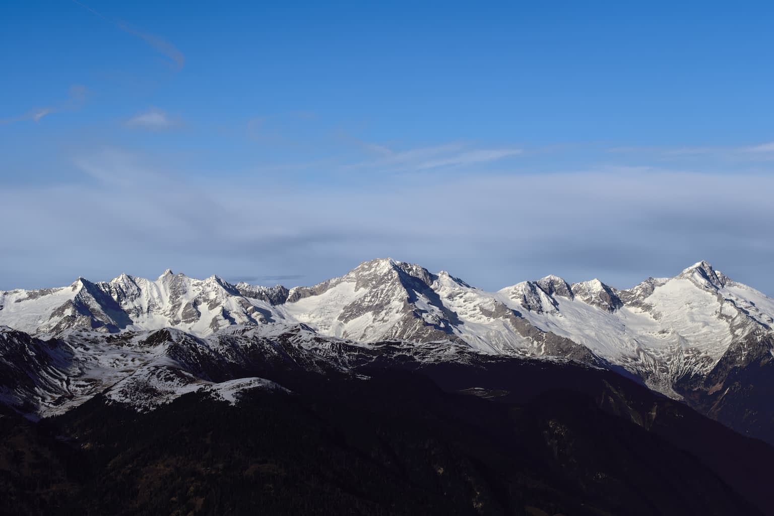 Monochrome image of the Schwarzenstein mountain (among Hornspitz, Großer Löffler, and others) as seen from the South, covered in snow with a layer of clouds and a blue sky above.