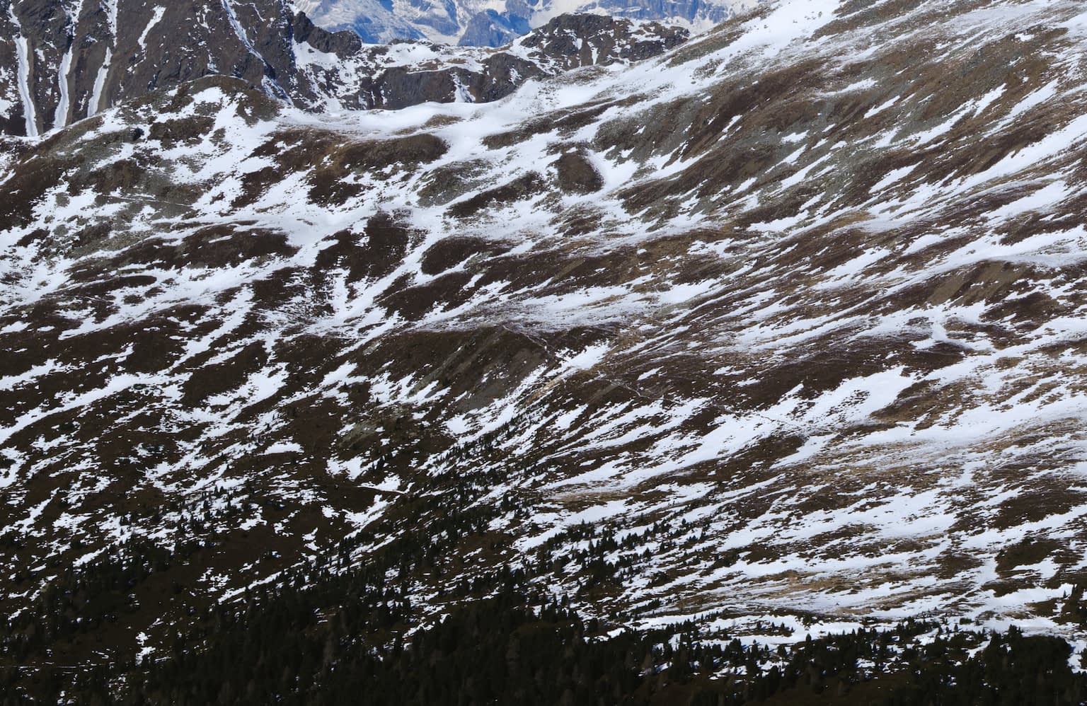 A view of the face of a mountain range looking painterly with brush strokes of white snow, brown earth, green trees, and blue mountain peeks. The view is from the Königsanger peek towards the combined east face of the Kaserbacher Hörndle and Märzenhörndle mountains in Schalders, South Tyrol. In the background are the Pflerscher Tribulaun and Gschnitzer Tribulaun peeks as well as the Schwarze Wand.