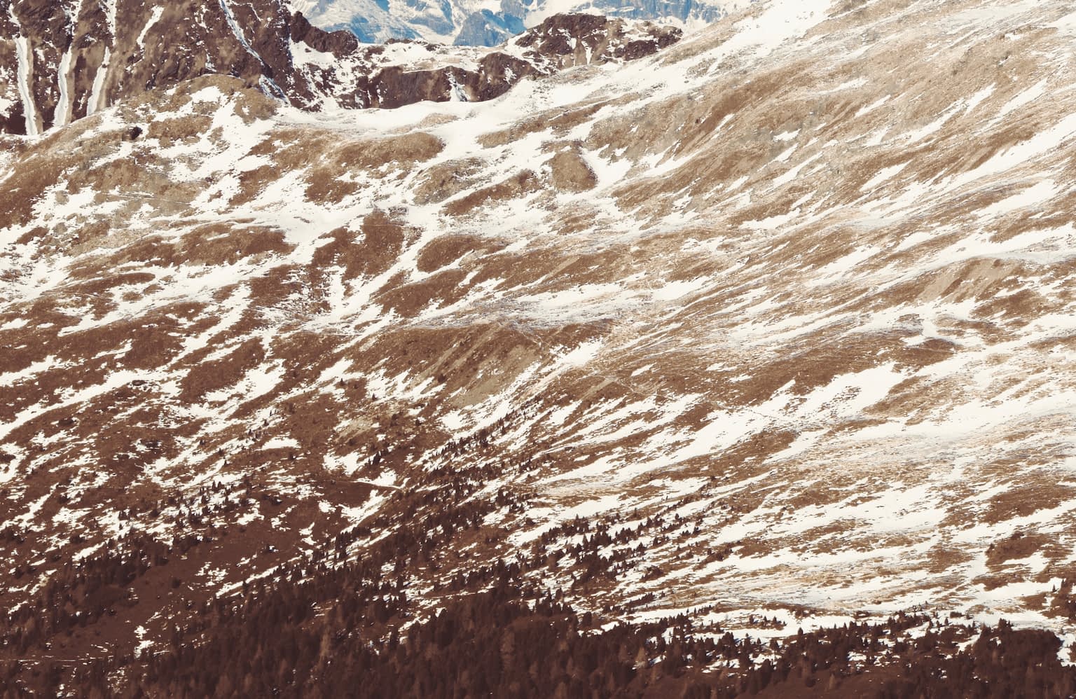 A view of the face of a mountain range looking painterly with brush strokes of white snow, brown earth, green trees, and blue mountain peeks. The view is from the Königsanger peek towards the combined east face of the Kaserbacher Hörndle and Märzenhörndle mountains in Schalders, South Tyrol. In the background are the Pflerscher Tribulaun and Gschnitzer Tribulaun peeks as well as the Schwarze Wand.
