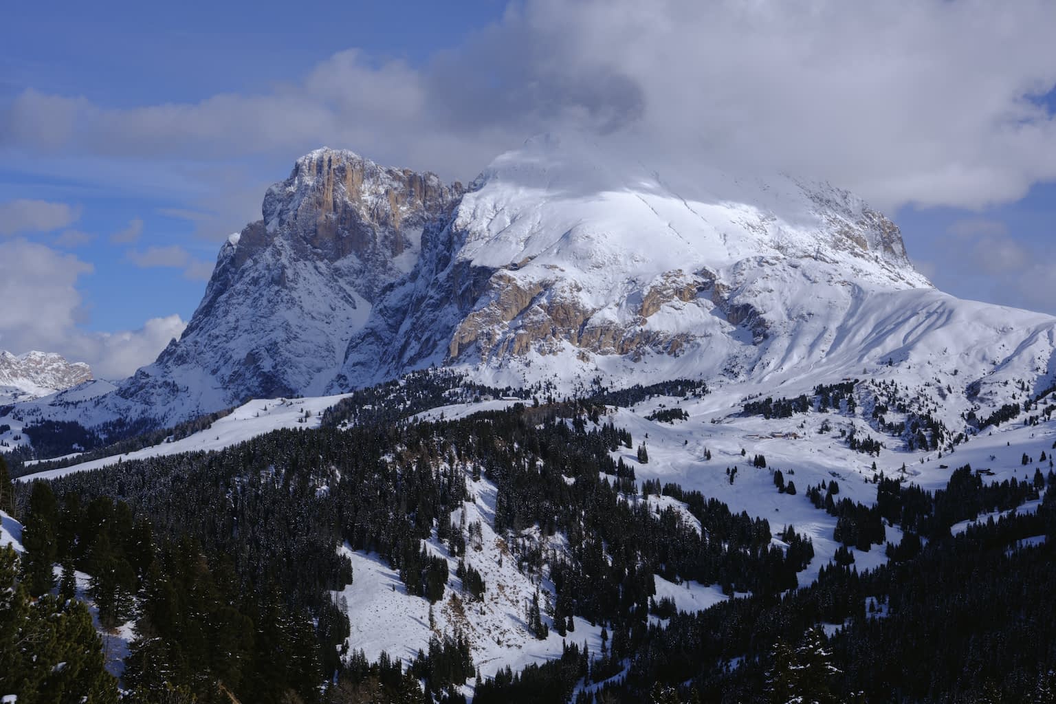 The Langkofel mountain range prominently featuring the Plattkofel covered in snow below and by a cloud from above on an otherwise sunny winter day.