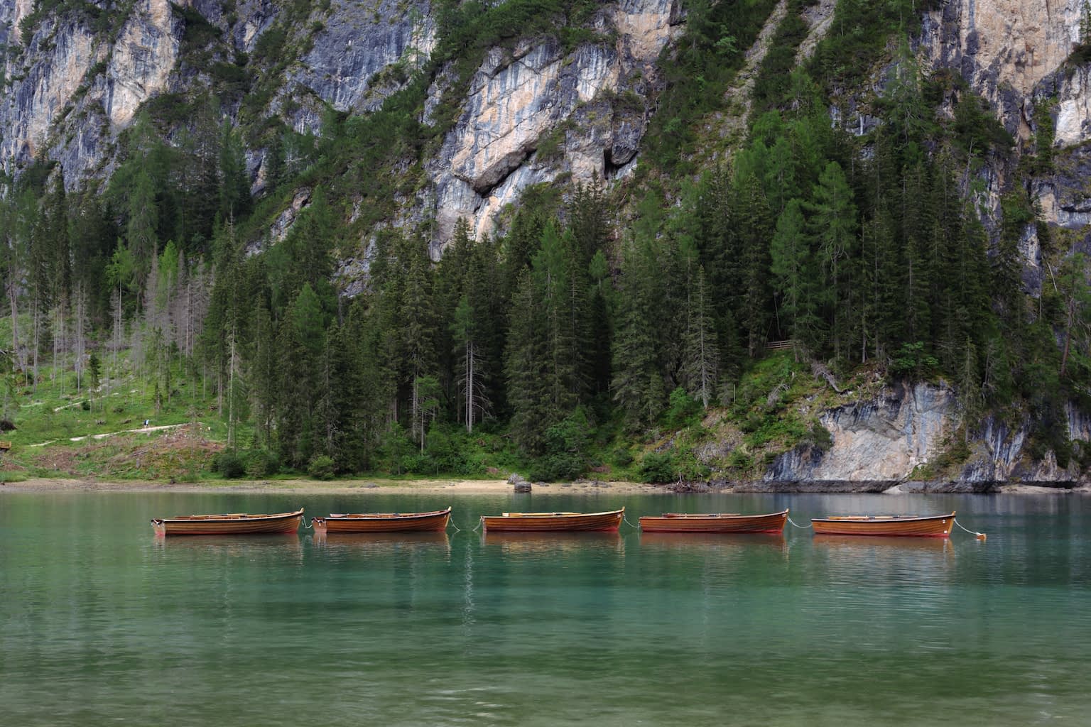 Five wooden rowing boats linked by ropes in the Pragser Wildsee with blue-green water and a steep rock face with trees rising in the background.