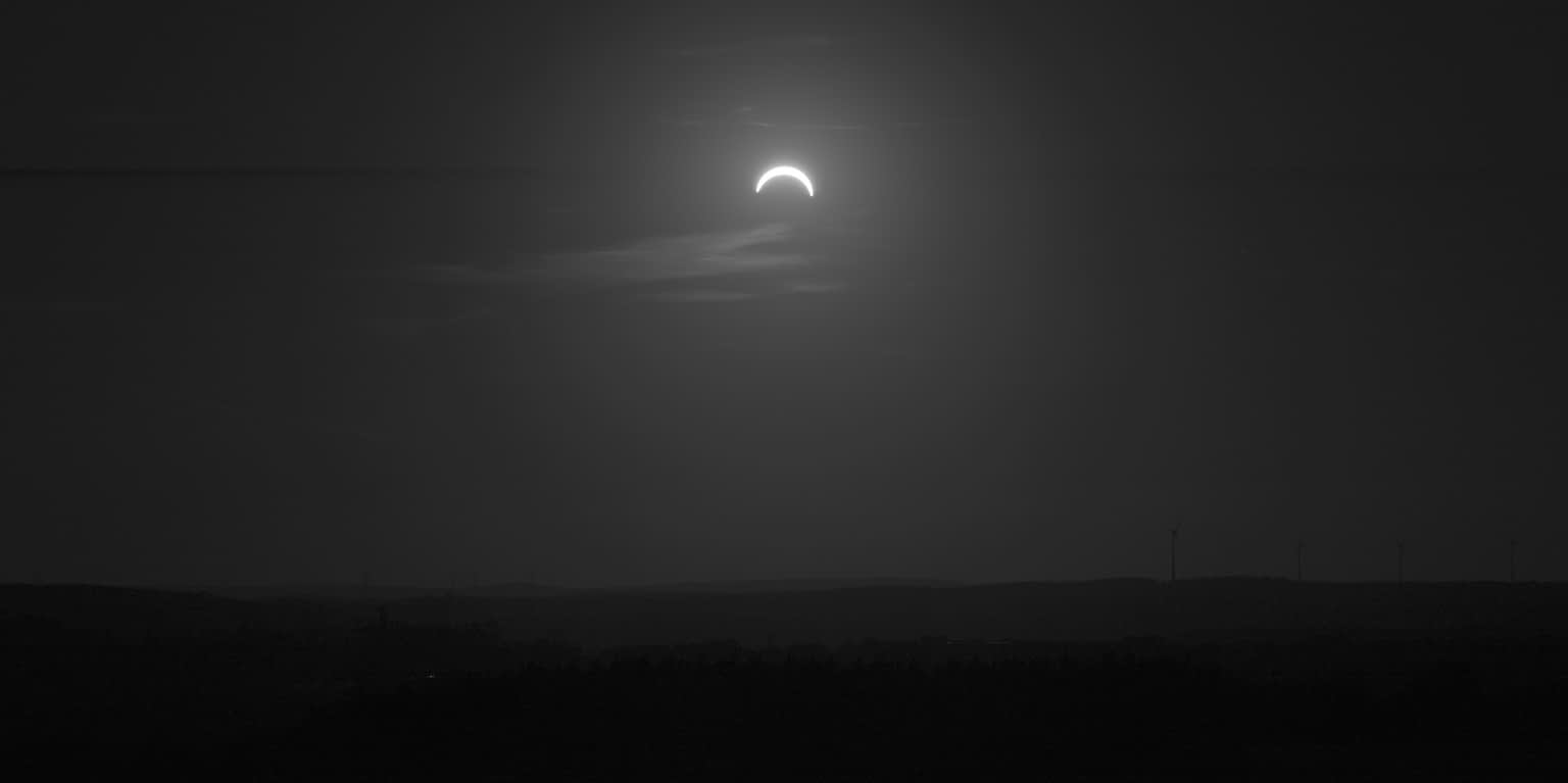Grayscale photo of a partial eclipse over a field with wind turbines.